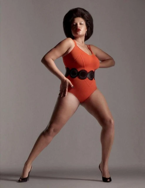 A woman with dark, voluminous hair poses in an orange bathing suit and black high heels against a neutral backdrop in a studio shot.