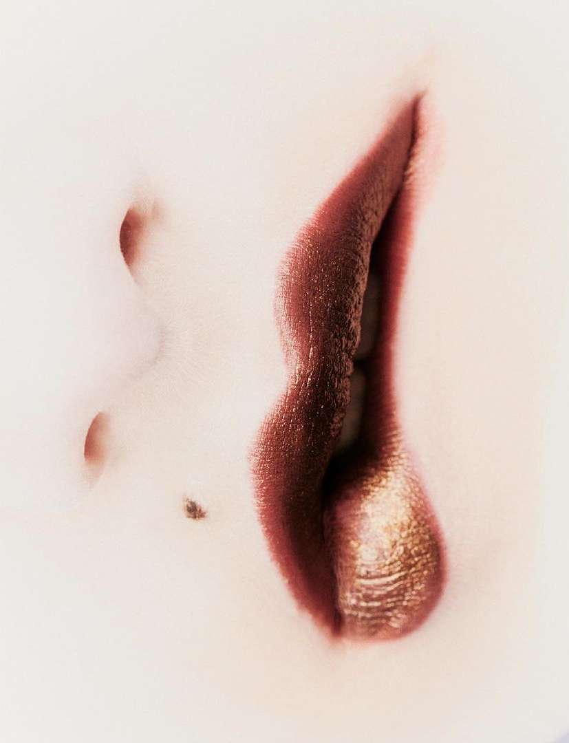 Close-up of person's face featuring lips with bronze-colored lipstick and a mole, set against a light-toned, out-of-focus background.