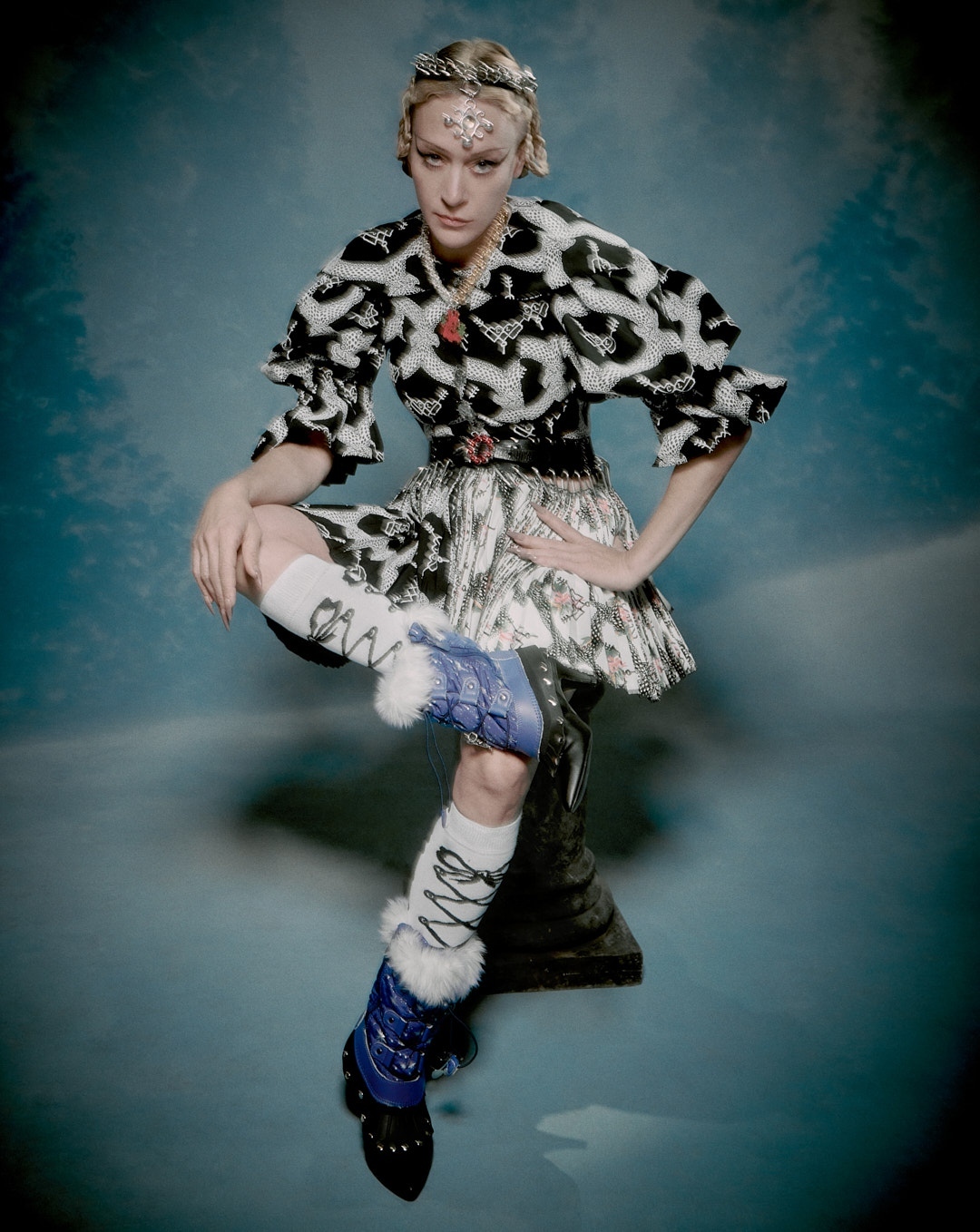 Woman poses in a black and white patterned dress with knee-high boots and decorative elements against a blue and cloudy backdrop.
