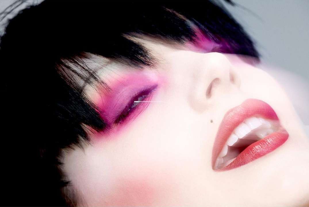 Close-up of a woman's face with pink eyeshadow and red lipstick, lying down with her head tilted back, highlighting her beauty features.