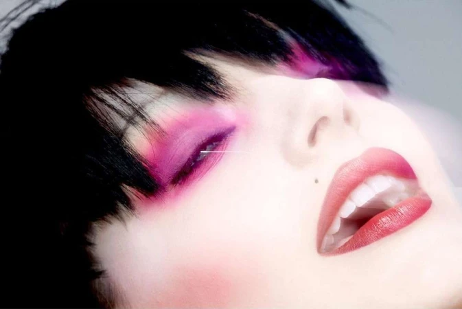 Close-up of a woman's face with pink eyeshadow and red lipstick, lying down with her head tilted back, highlighting her beauty features.
