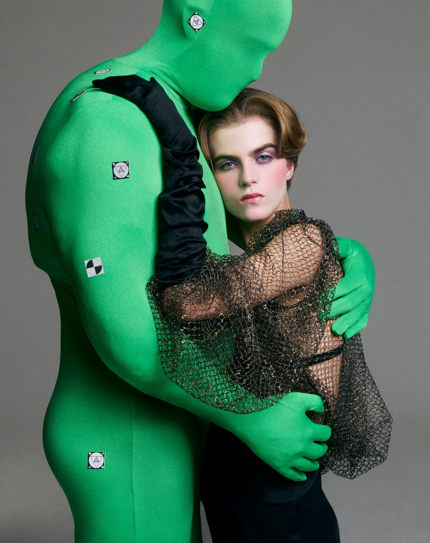 A fair-skinned woman with dark hair and makeup hugs a green-suited figure with tracking markers against a gray background in a studio setting.