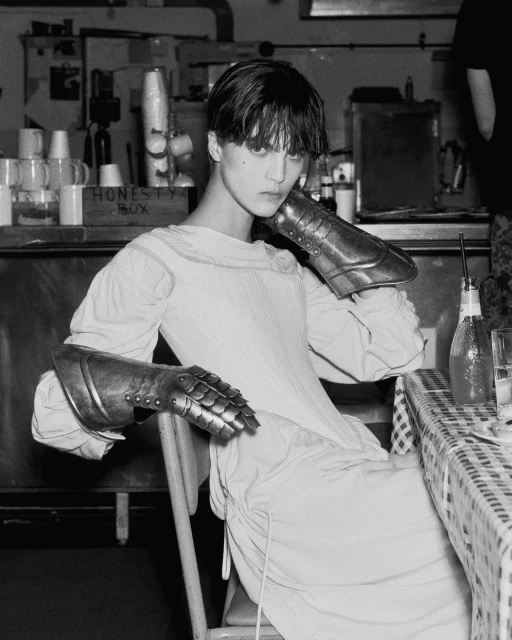 Monochrome portrait of a person seated, wearing medieval armor gauntlets and a pale dress, sitting at a checkered table indoors.