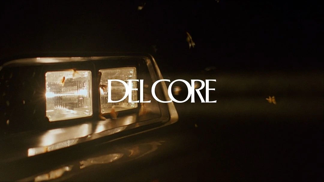 Close-up of a vintage car headlight, surrounded by moths at night, illuminated by a warm glow with white Delcore text overlay.