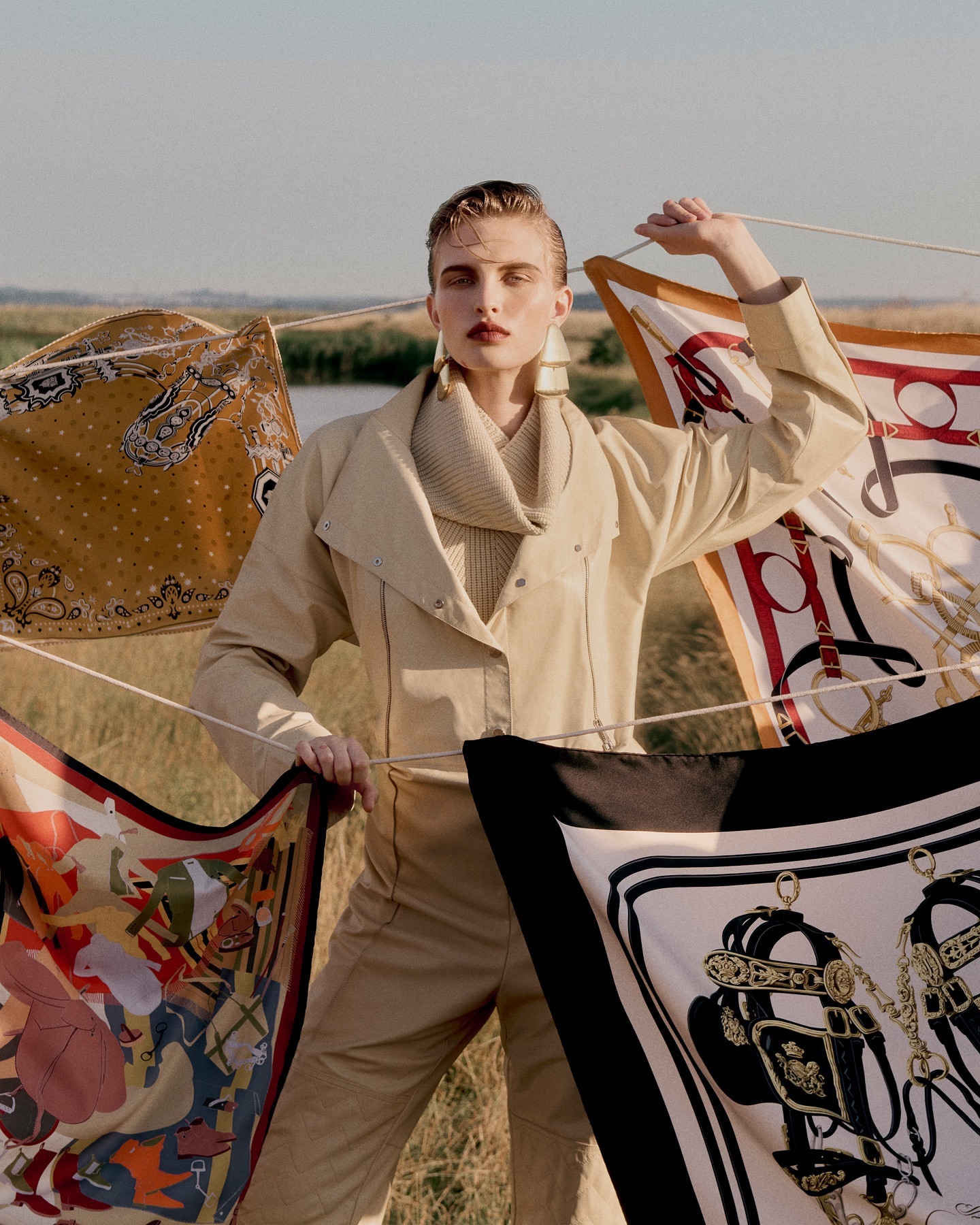 A young adult fashion model poses outdoors in a beige jacket and pants, surrounded by colorful silk scarves on a cloudy day.