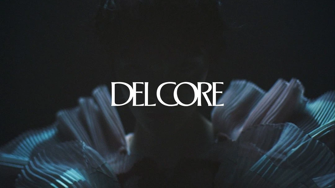 Closeup of a person with a ruffled, pleated garment in a dark, moody scene, with the text 'DELCORE' overlaid in white.