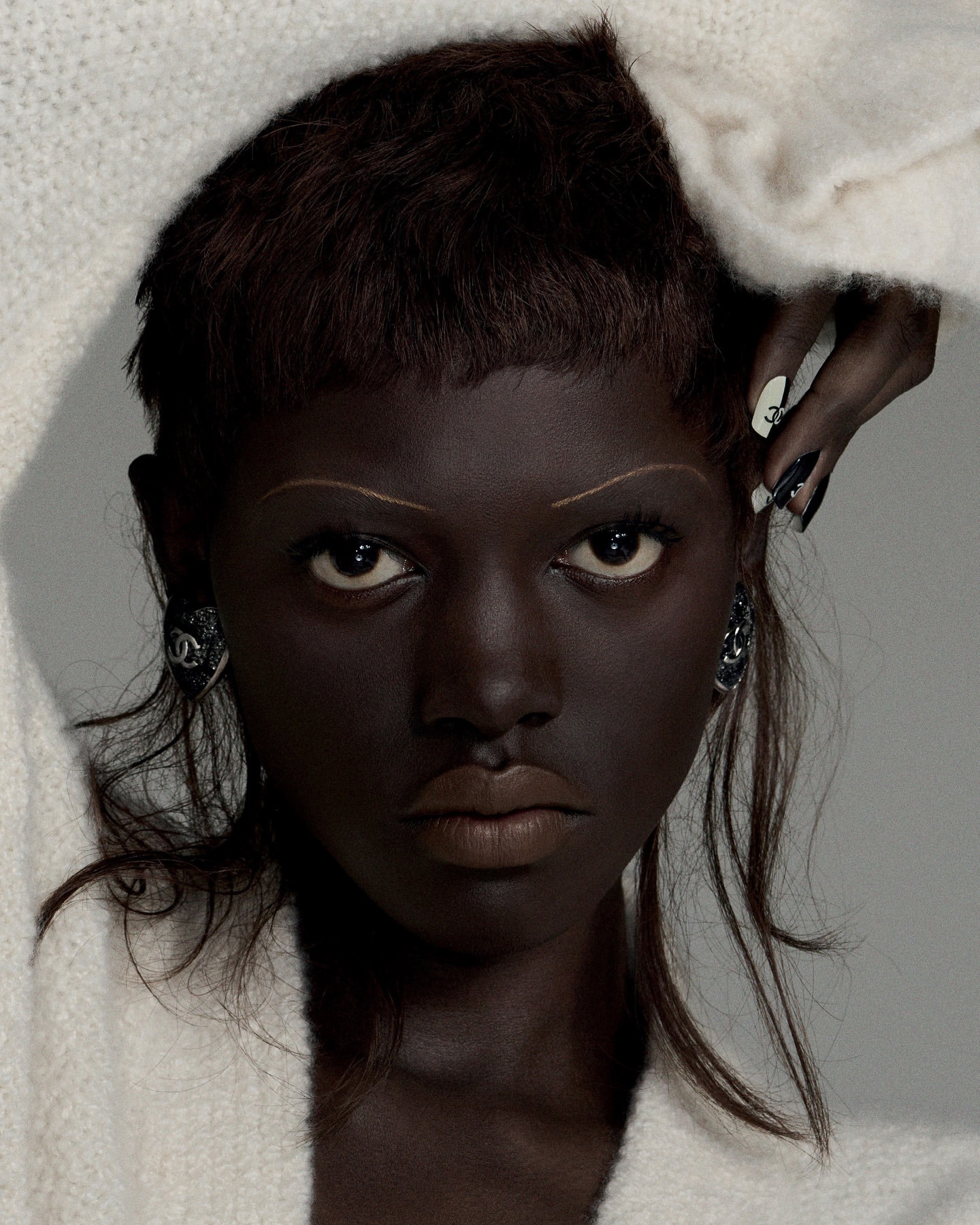 Close-up portrait of a young adult female with dark skin and a serious expression, adorned with distinctive makeup and jewelry.