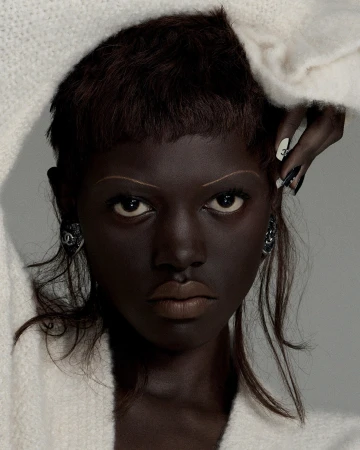 Close-up portrait of a young adult female with dark skin and a serious expression, adorned with distinctive makeup and jewelry.