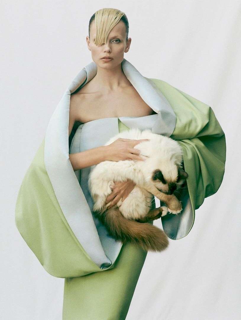 Blonde woman in couture mint green dress holding a fluffy white and brown cat on a plain white backdrop in a fashion photography style.