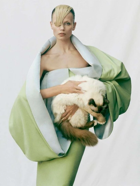 Blonde woman in couture mint green dress holding a fluffy white and brown cat on a plain white backdrop in a fashion photography style.