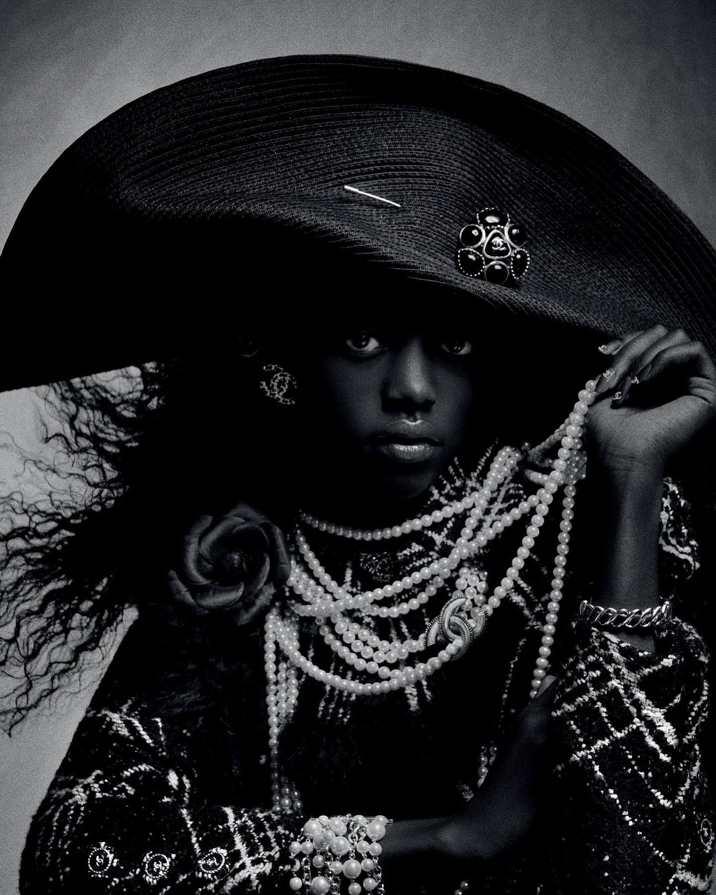 A solitary Black model wearing oversized black hat, tweed jacket, and pearls in a fashion-forward pose, against a studio backdrop.