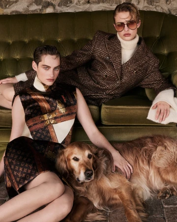 Two female models wearing luxury dresses pose with a golden retriever on an olive-green couch in front of a stone wall inside.