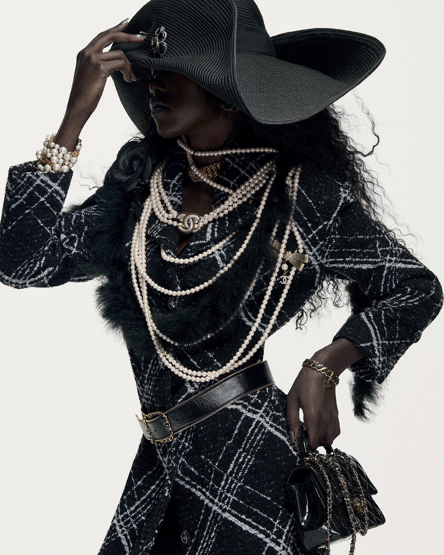 Dark-skinned woman in black tweed coat dress, wide-brim hat, pearls, and black bag, poses against a bright background in fashion shot.