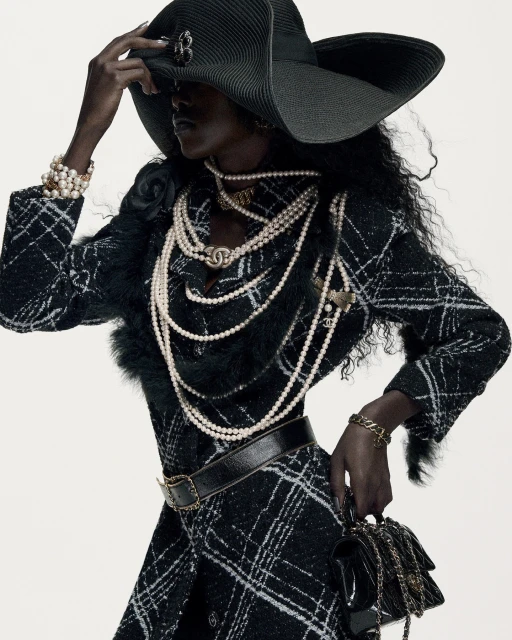 Dark-skinned woman in black tweed coat dress, wide-brim hat, pearls, and black bag, poses against a bright background in fashion shot.