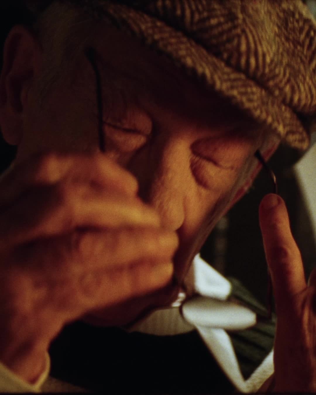 Close-up of an elderly adult male wearing a tweed cap, holding glasses and a thin wire, indoors with a dark orange color grade.