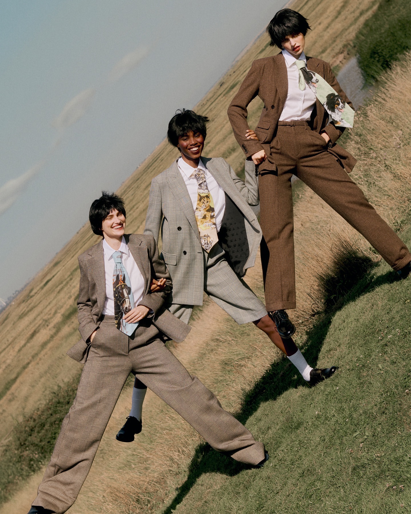 Three women in stylish suits and ties pose outdoors in a grassy field under a bright sky, showcasing fashion and elegance.