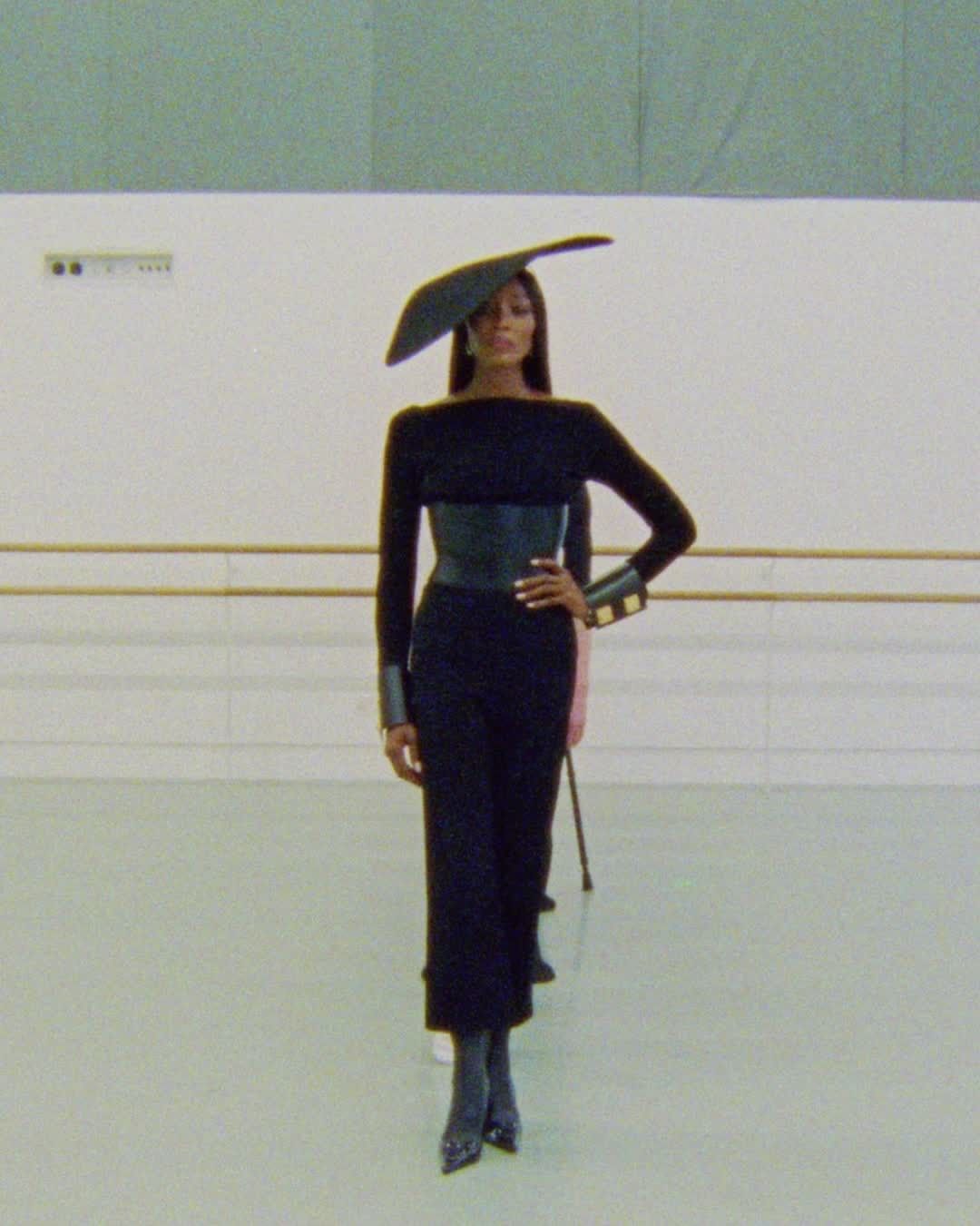 A woman models a black outfit and large hat on a runway. She has on a belt, cuffs and heels, and is posing with her hand on her hip.