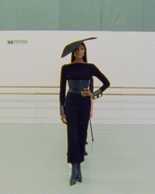 A woman models a black outfit and large hat on a runway. She has on a belt, cuffs and heels, and is posing with her hand on her hip.