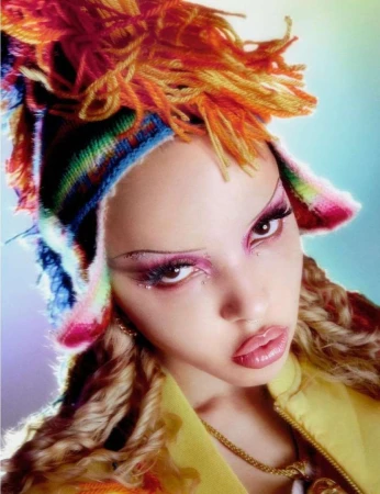 Close-up of a woman with dramatic makeup and a colorful hat, wearing a yellow jacket and jewelry against a bright, soft background.