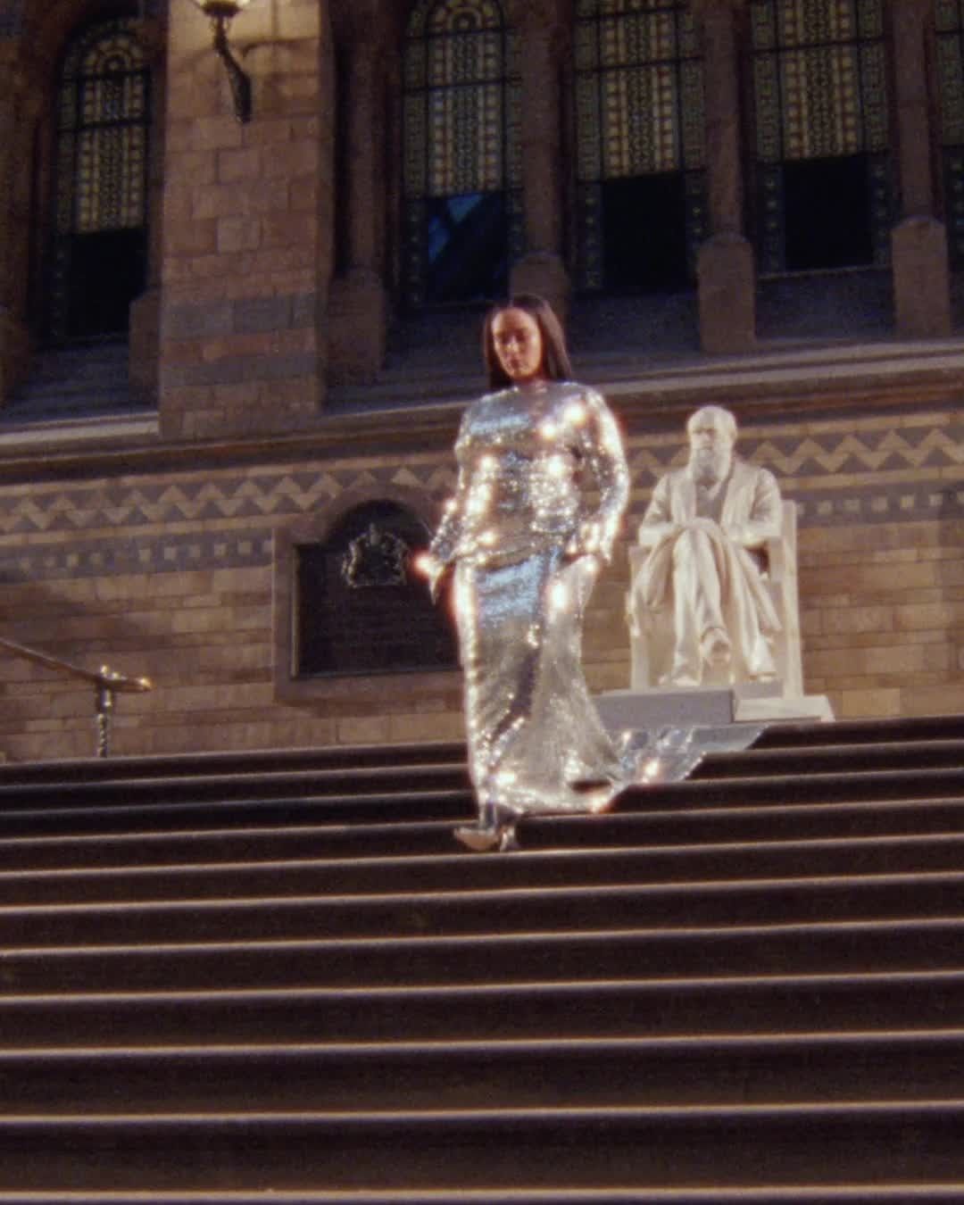 Woman in sequin gown descends stairs of stone building; sculpture on landing; day exterior shot; fashion, architecture, travel.