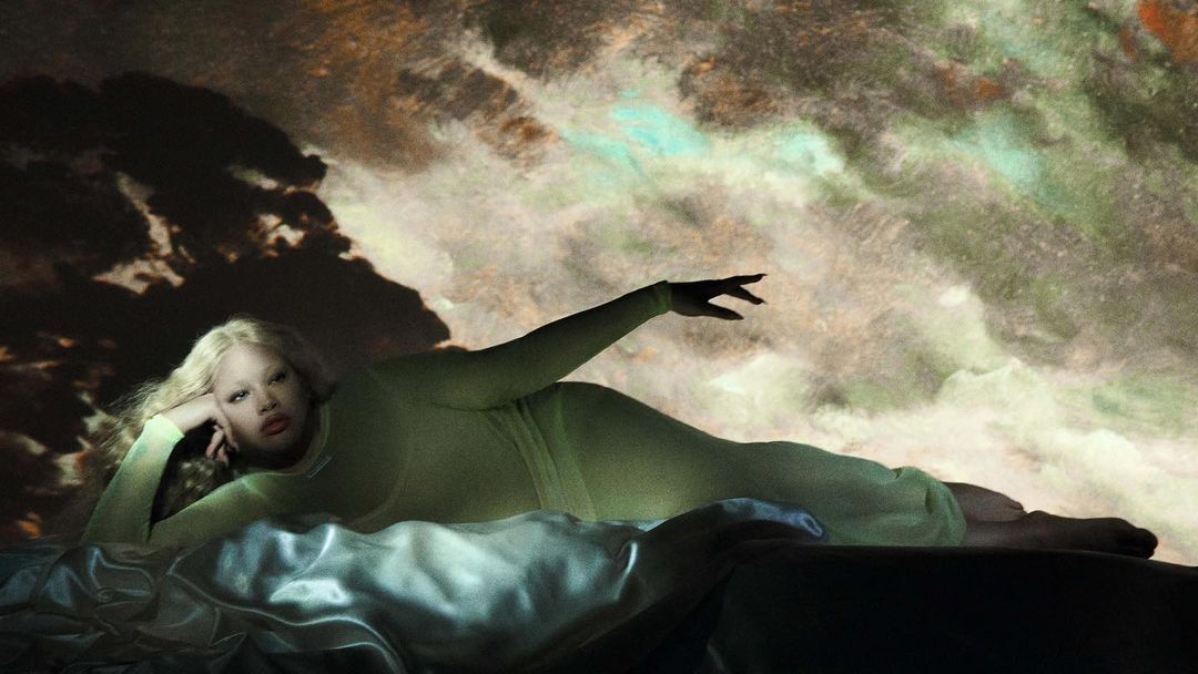 Woman with long blonde hair lying on bed with an arm raised in a studio. A textured cloudy background is behind the woman.