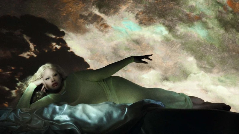 Woman with long blonde hair lying on bed with an arm raised in a studio. A textured cloudy background is behind the woman.