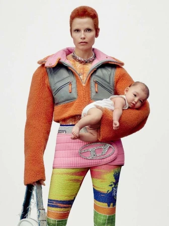 Woman with short red hair holds a baby in a white diaper. She wears an orange jacket, pink skirt with a Diesel logo, and patterned leggings.