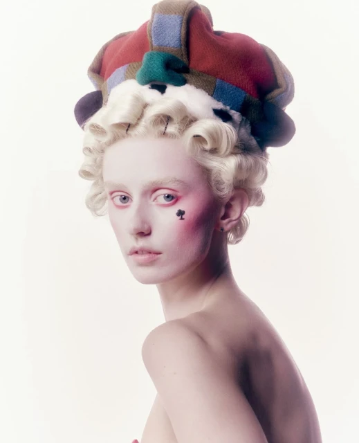 Elegant woman with pale skin and elaborate hairstyle, wearing a colorful hat and spade makeup, posing against a plain background.