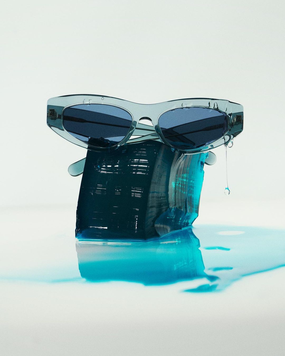 Stylish translucent blue sunglasses perched on a rough block of blue resin, with light reflecting on the wet white surface below.