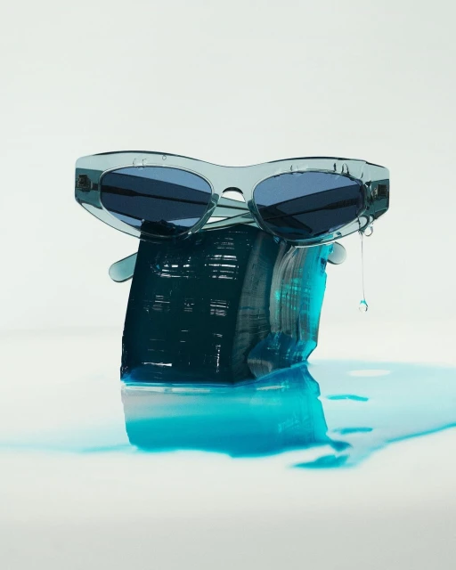 Stylish translucent blue sunglasses perched on a rough block of blue resin, with light reflecting on the wet white surface below.