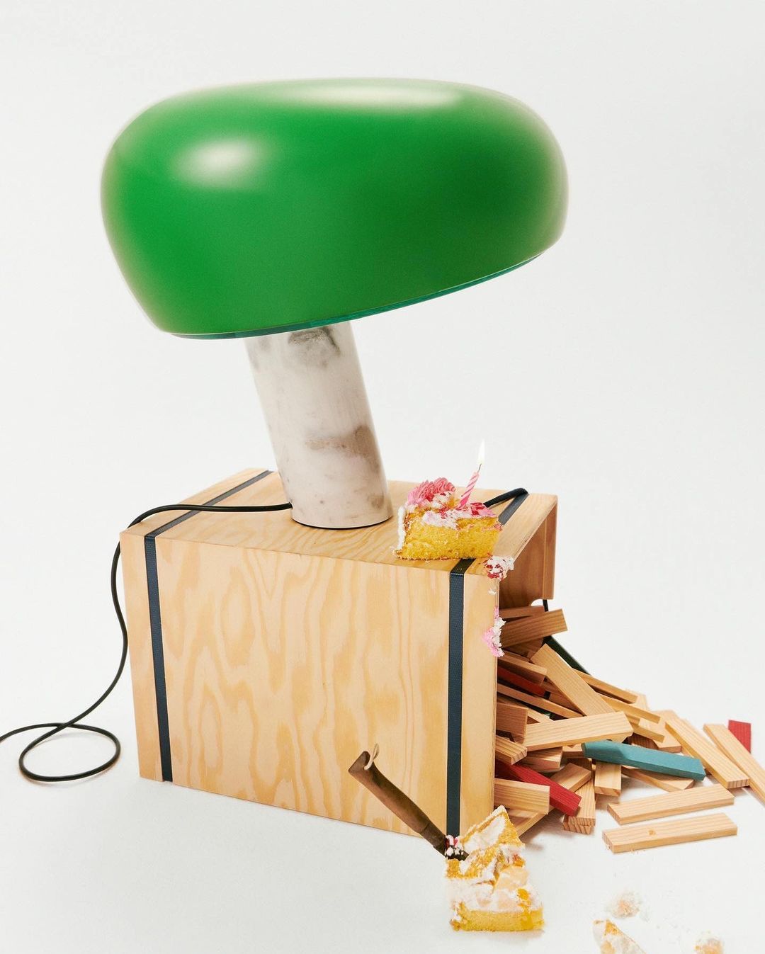 A playful product shot features a green mushroom lamp atop a wooden crate with scattered wooden blocks and a slice of birthday cake.
