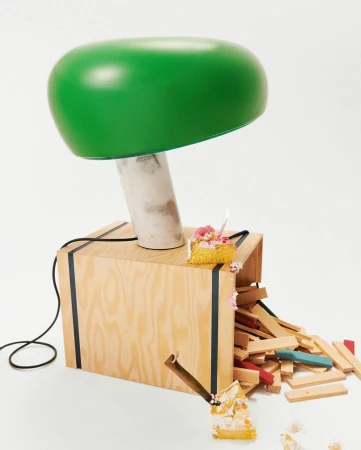 A playful product shot features a green mushroom lamp atop a wooden crate with scattered wooden blocks and a slice of birthday cake.