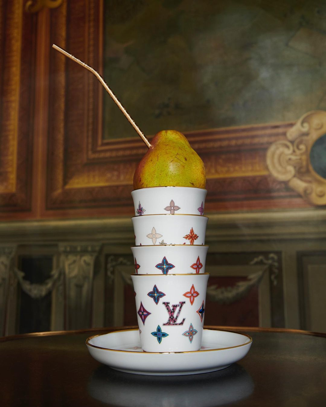 Stacked designer cups and pear on table against a wall with a painting in a bright indoor setting, providing a unique still-life composition.