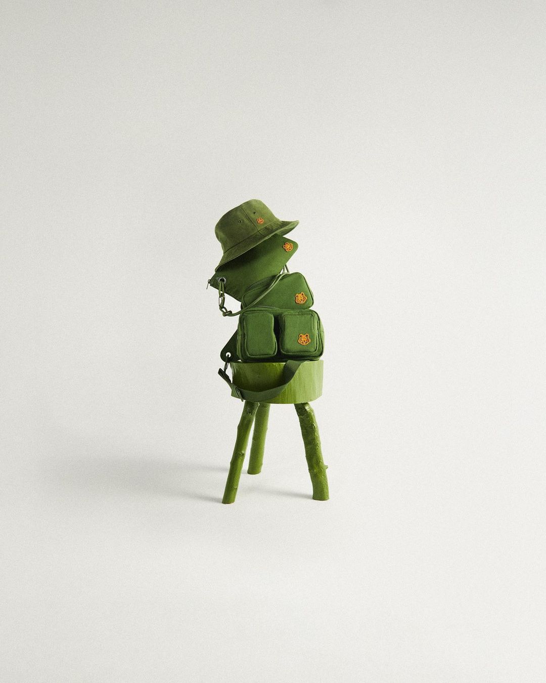 Still life shot of a green bucket hat and two small bags stacked on a green faux-wood stand against a light grey background.