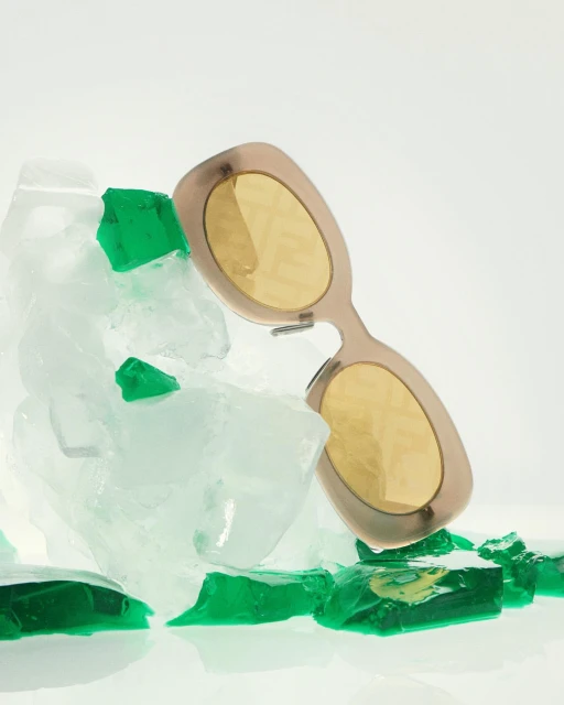 Sunglasses with beige frames and yellow lenses rest on clear and green crushed ice in a studio setting with a white background.