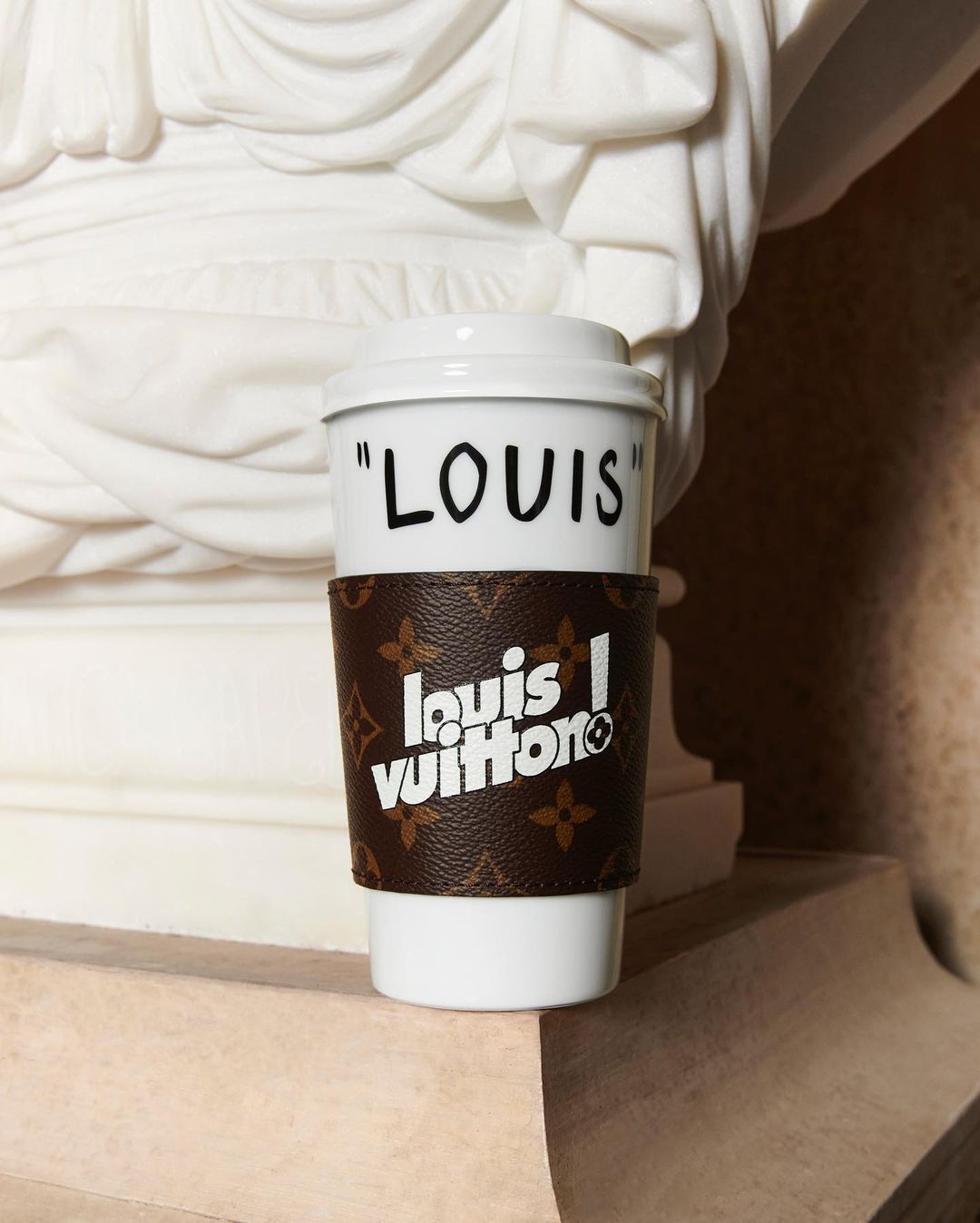 White travel cup with lid and Louis Vuitton sleeve sits on marble surface in front of neoclassical marble sculpture.