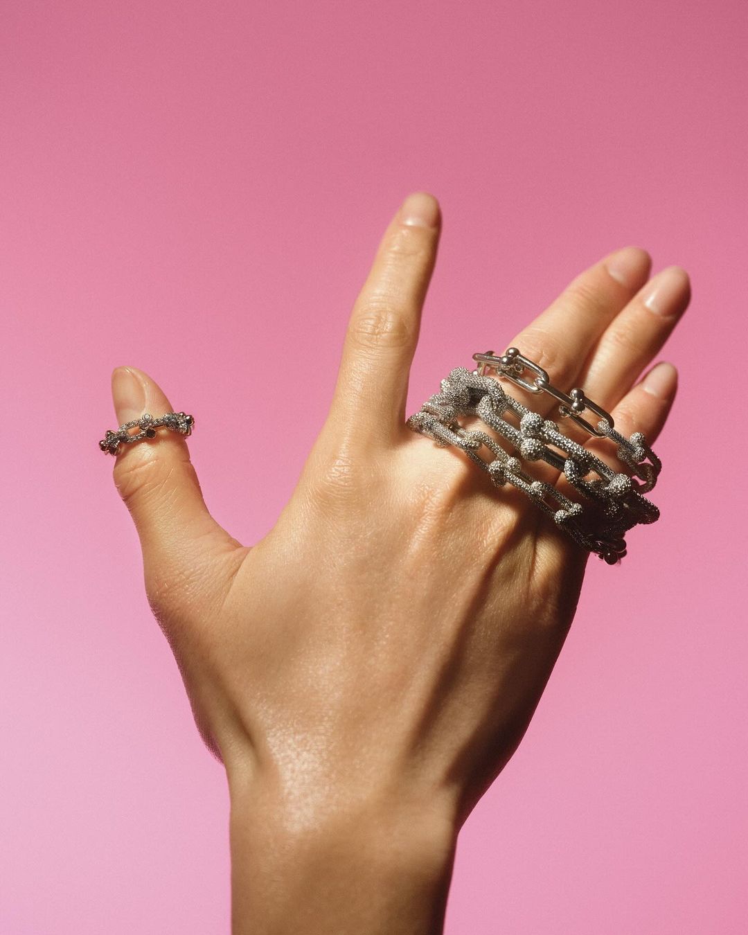 Close-up studio shot of a fair-skinned adult hand displaying silver metal jewelry: a ring and multiple chain bracelets, pink background.