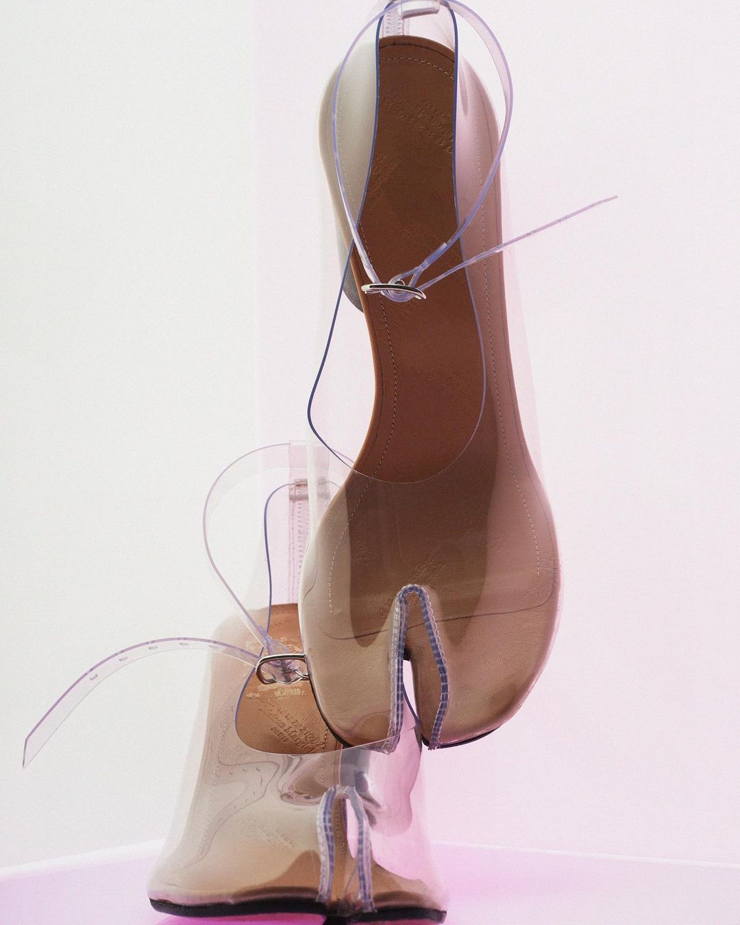 Split-toe sandals with clear straps and block heels on a pink surface against a white background, highlighting a modern and unique design.