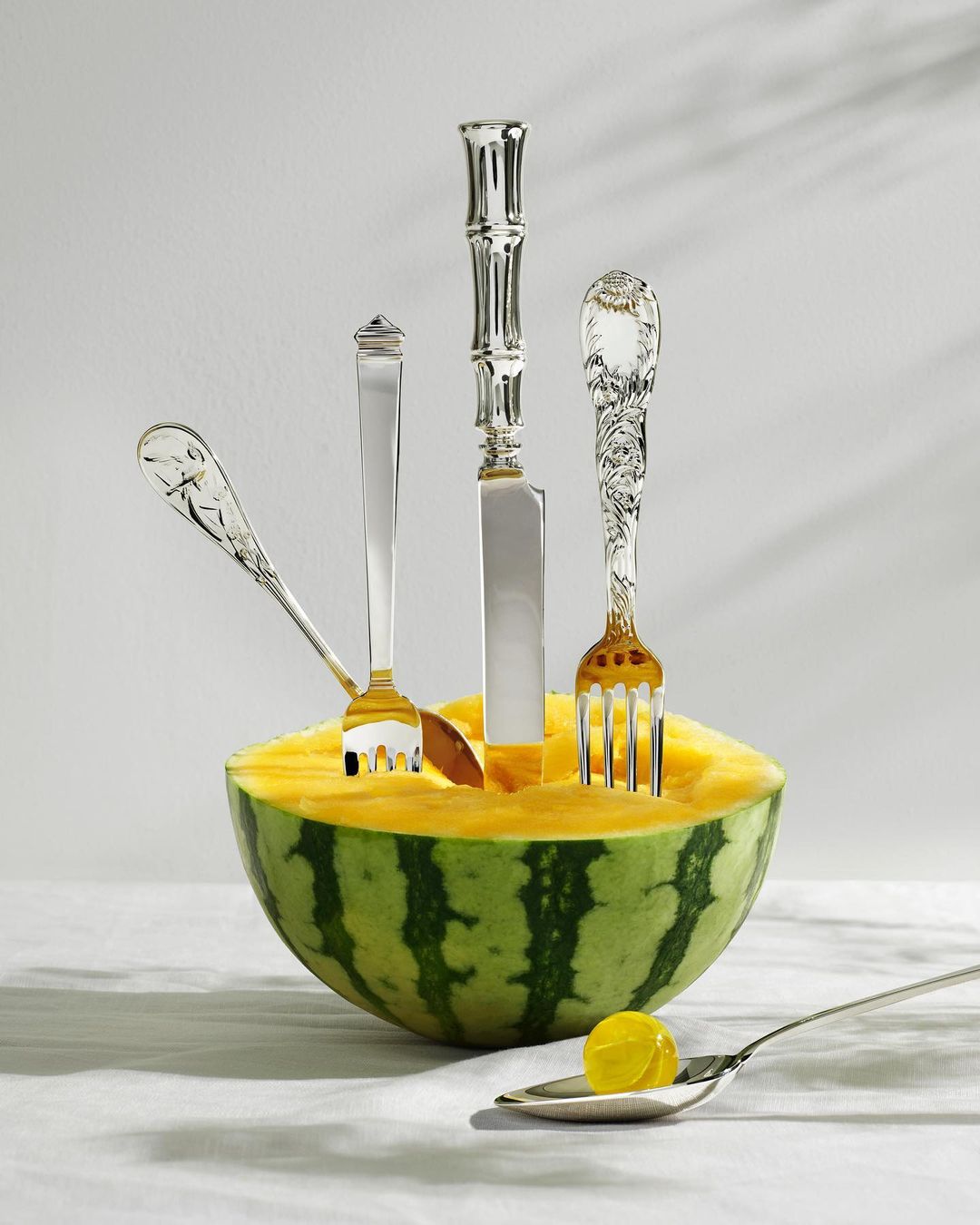 A yellow watermelon is creatively displayed with silver forks and a knife stuck into it; a small yellow fruit sits on a spoon nearby.