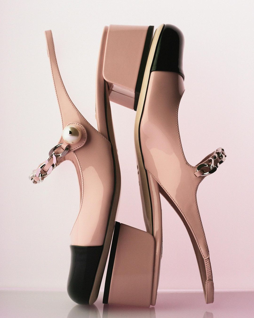 Elegant fashion product shot featuring a pair of light-pink patent leather heeled shoes with black toe caps and silver chain detail.