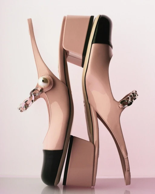 Elegant fashion product shot featuring a pair of light-pink patent leather heeled shoes with black toe caps and silver chain detail.