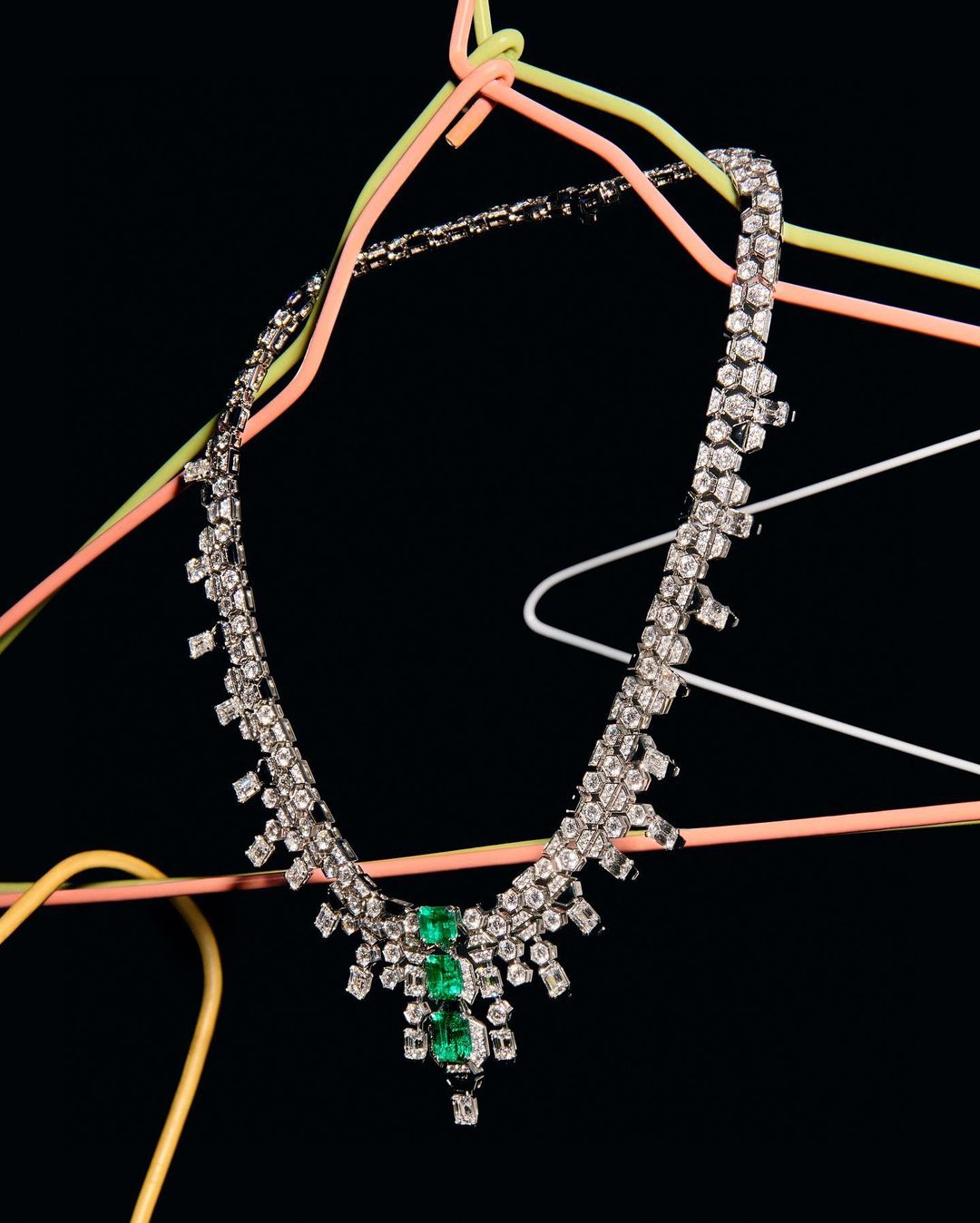 Elegant emerald and diamond necklace on colorful clothes hangers against a stark black backdrop, jewelry photography, high-contrast lighting.