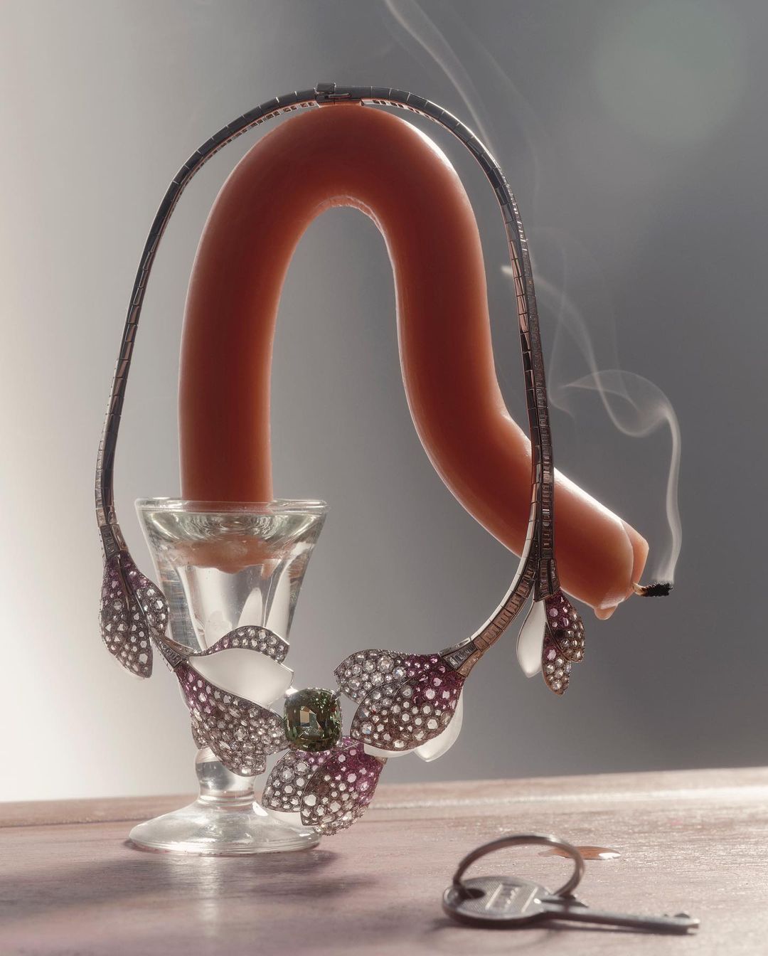 Fine jewelry necklace with flower designs displayed around a glass vase holding a sausage, on a surface with a key.