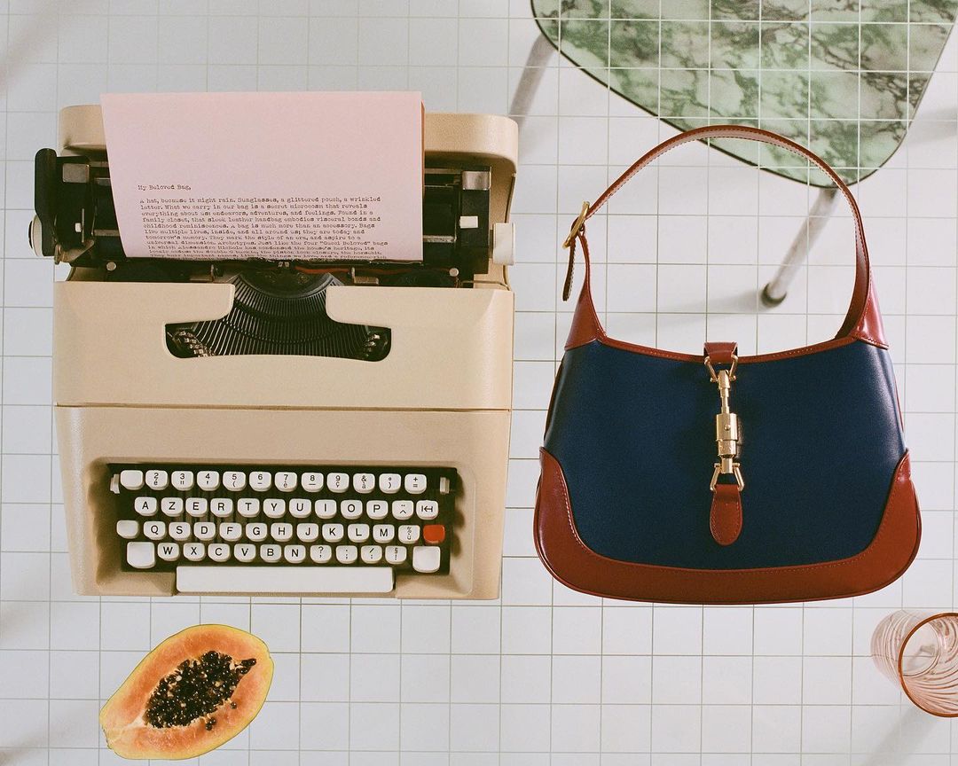Beige typewriter with pink paper next to a blue and brown leather handbag on a white tiled surface, with a green marble table leg.