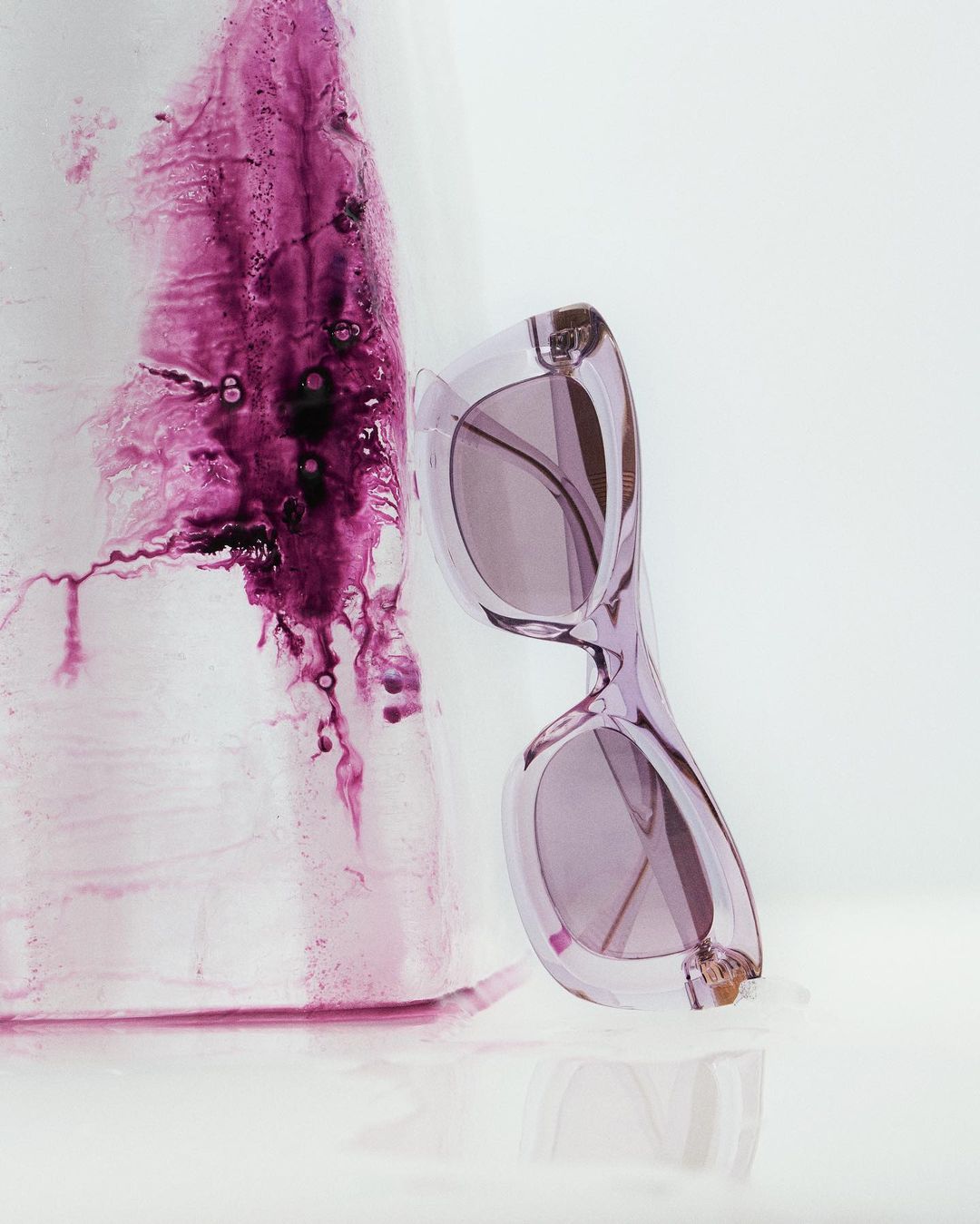 Stylish purple sunglasses next to an abstract artistic composition on a reflective white surface, creating a visually striking image.