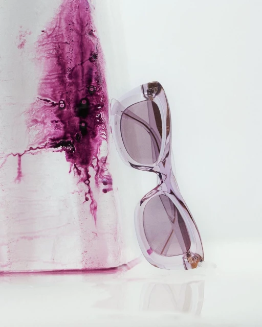 Stylish purple sunglasses next to an abstract artistic composition on a reflective white surface, creating a visually striking image.