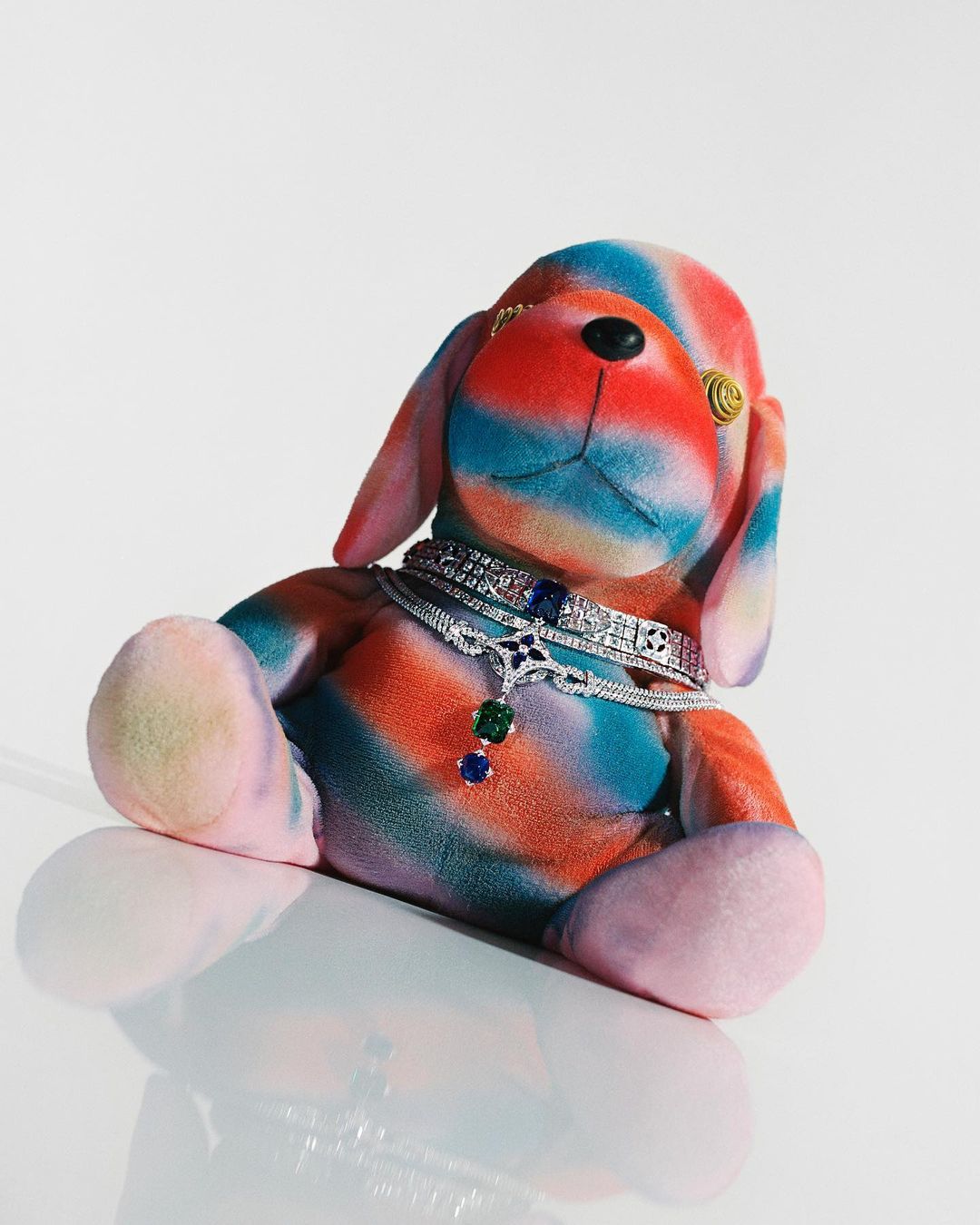 Tie-dye dog plush wearing diamond and sapphire jewellery sitting on a white reflective surface, creating a visually striking still life.