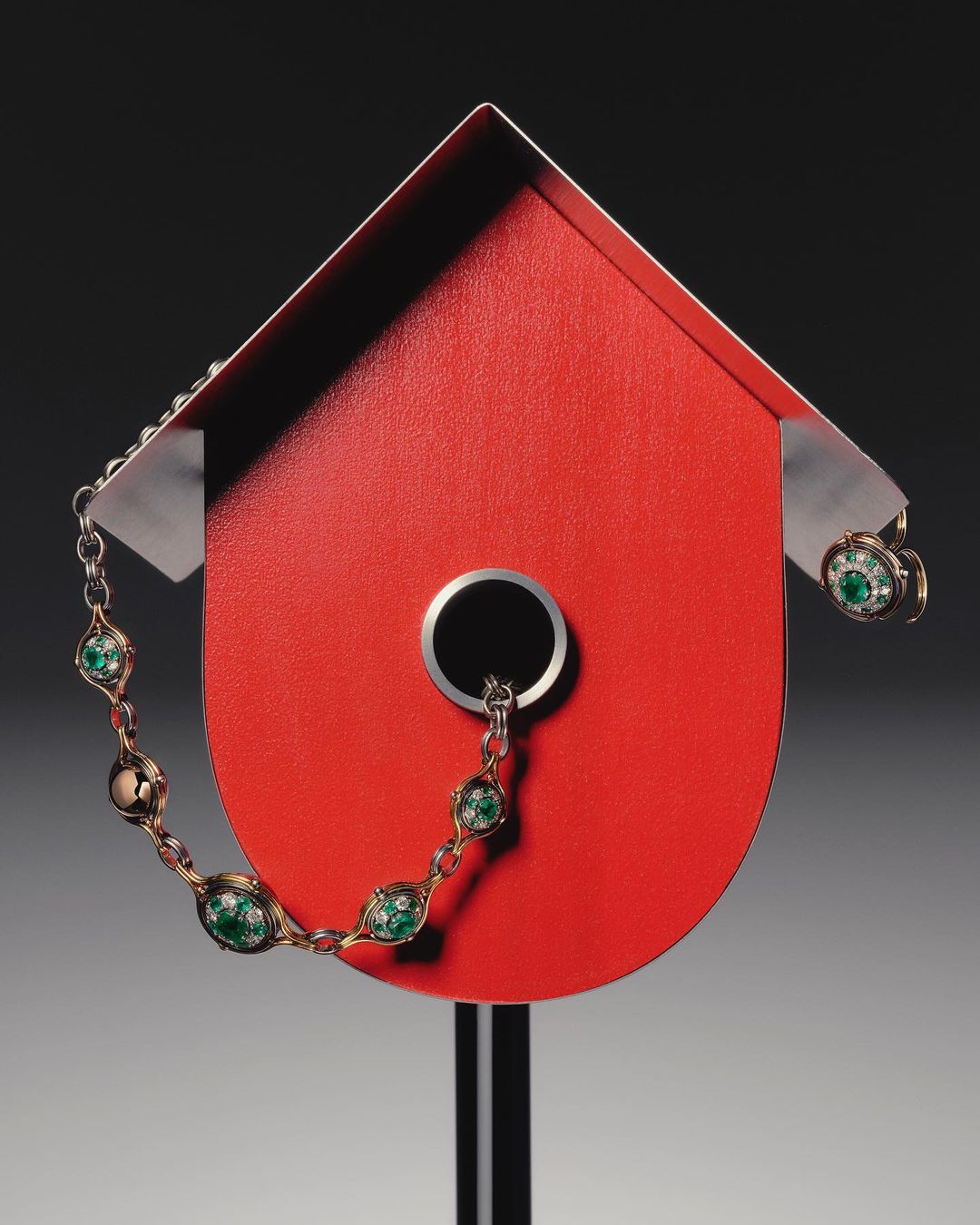 A red birdhouse on a gray background, adorned with a delicate, decorative necklace and earring set, creating a striking still life composition.