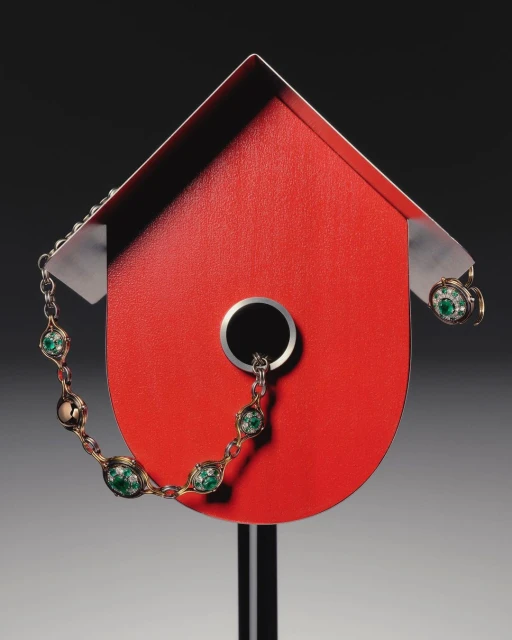 A red birdhouse on a gray background, adorned with a delicate, decorative necklace and earring set, creating a striking still life composition.