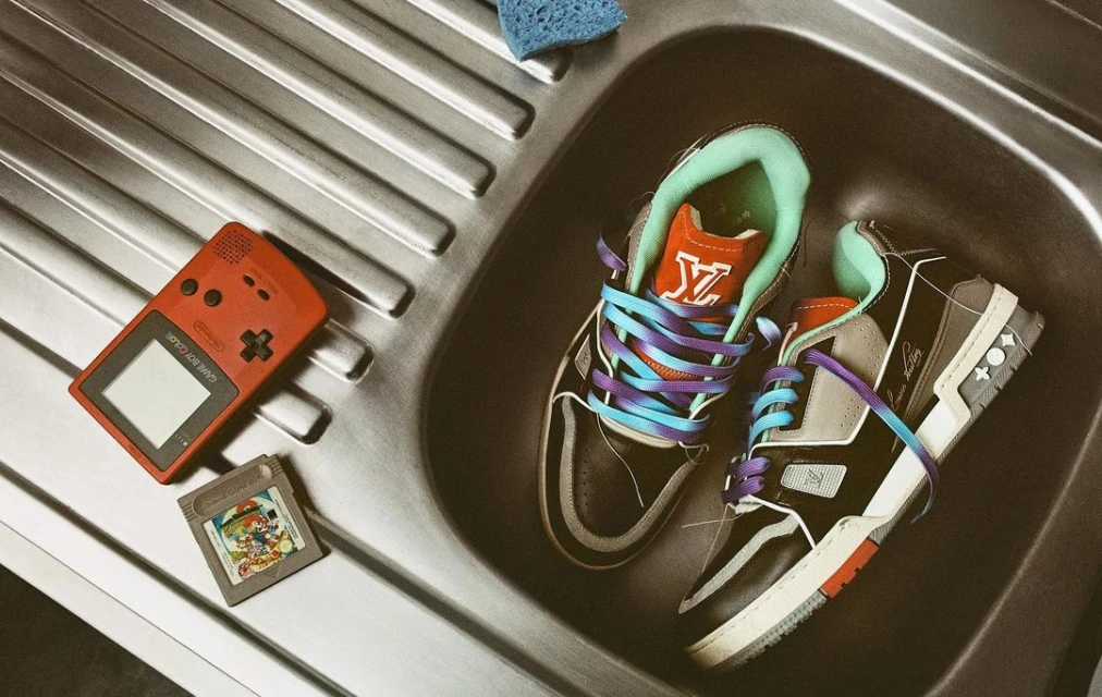 Two Louis Vuitton sneakers and a Game Boy in a silver sink with a blue sponge and a game cartridge for a stylish, retro gamer image.